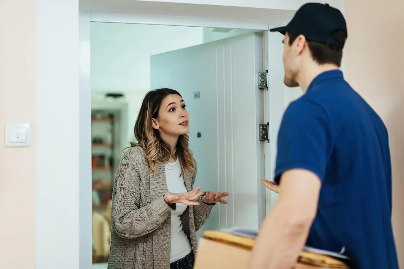 Молодая недовольная женщина стоит в дверях и разговаривает с курьером, принимая посылку.